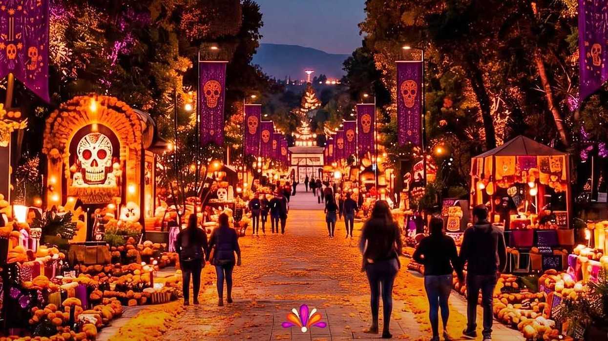 A candlelit Día de los Muertos festival fills a tree-lined walkway with skull banners, altars, pumpkins, and visitors.