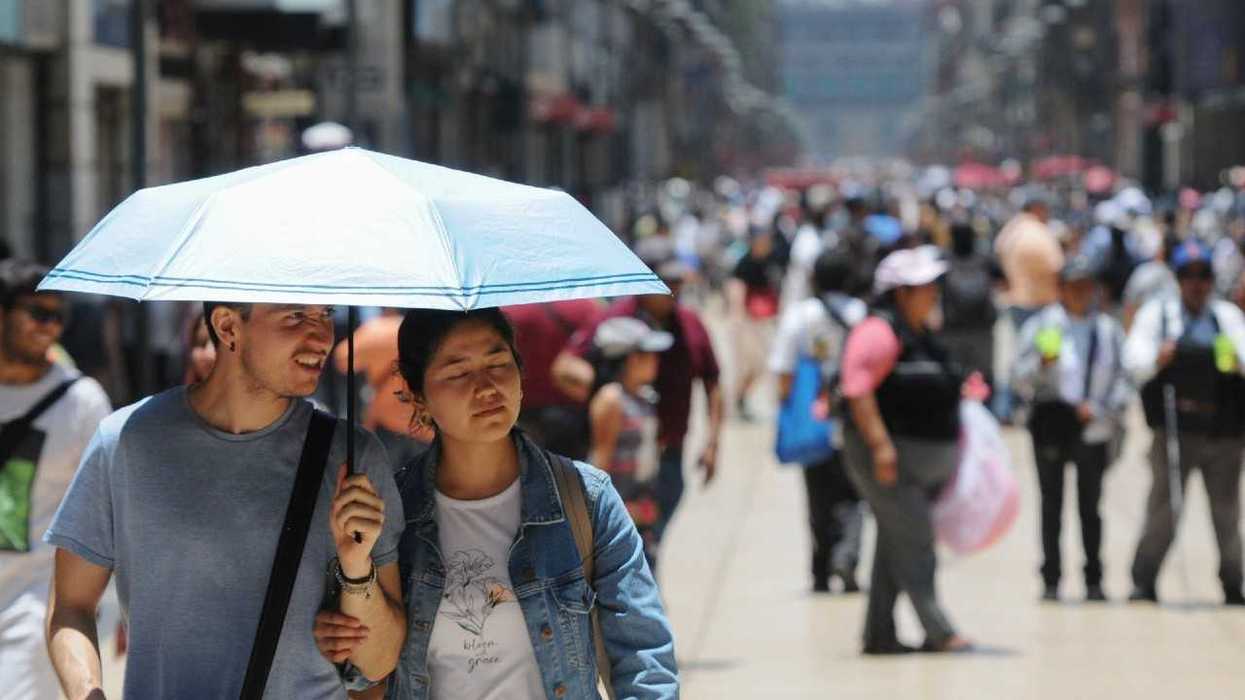 A couple shelters under a light blue umbrella amid a crowded, sunlit city street.