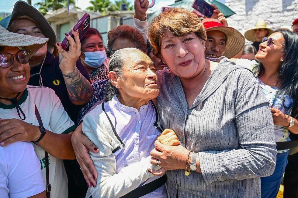 A group of smiling people, two women in front embracing joyfully outdoors.