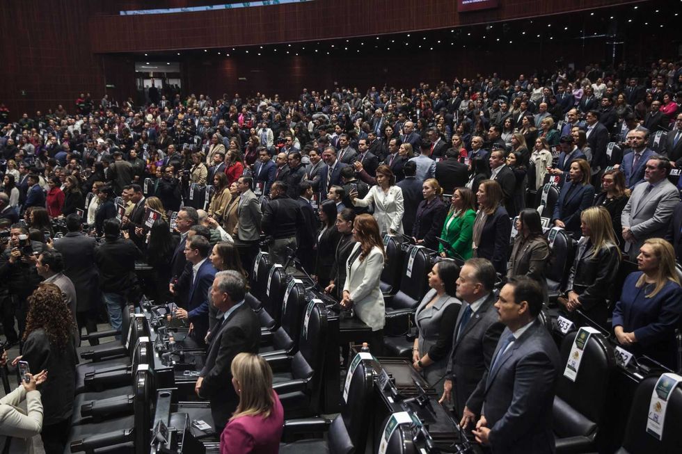 A large assembly of people stand in rows inside a legislative chamber.