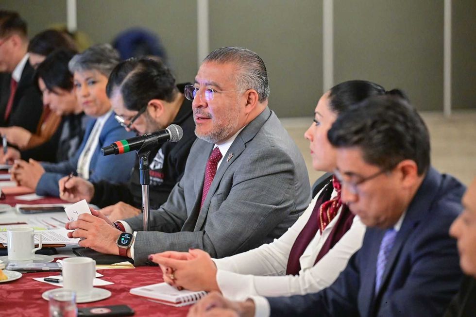 A man in a suit speaks into a microphone at a conference table with others.