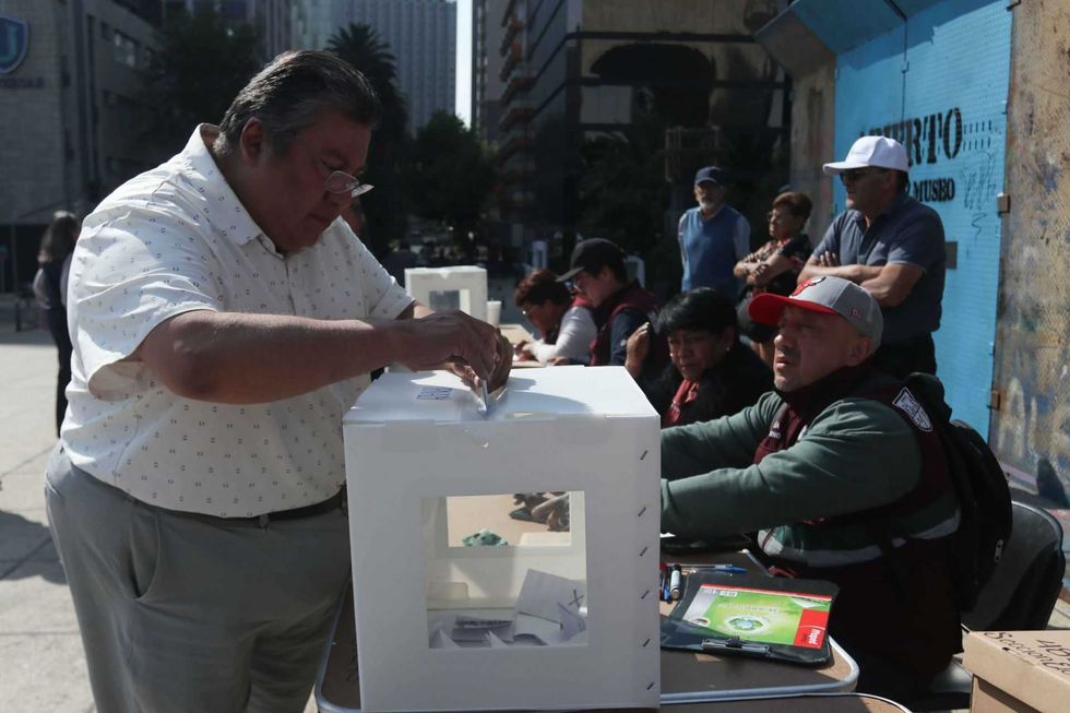 A person voting outdoors at a booth while others sit nearby and observe.