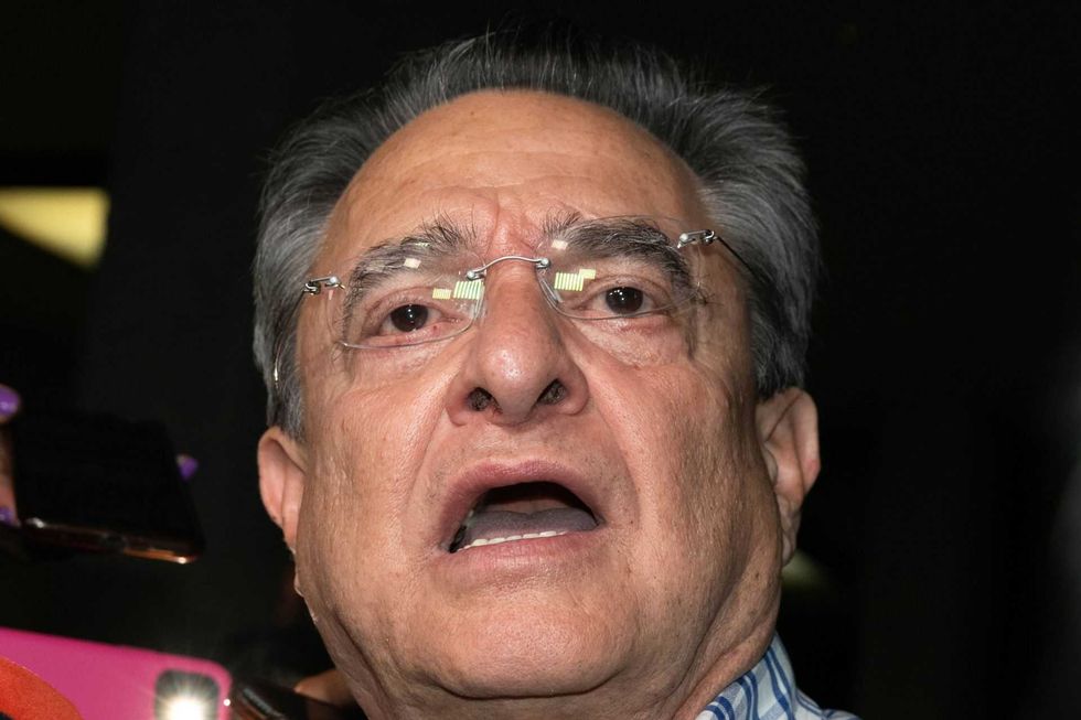 A surprised elderly man with glasses and open mouth, against a dark background.