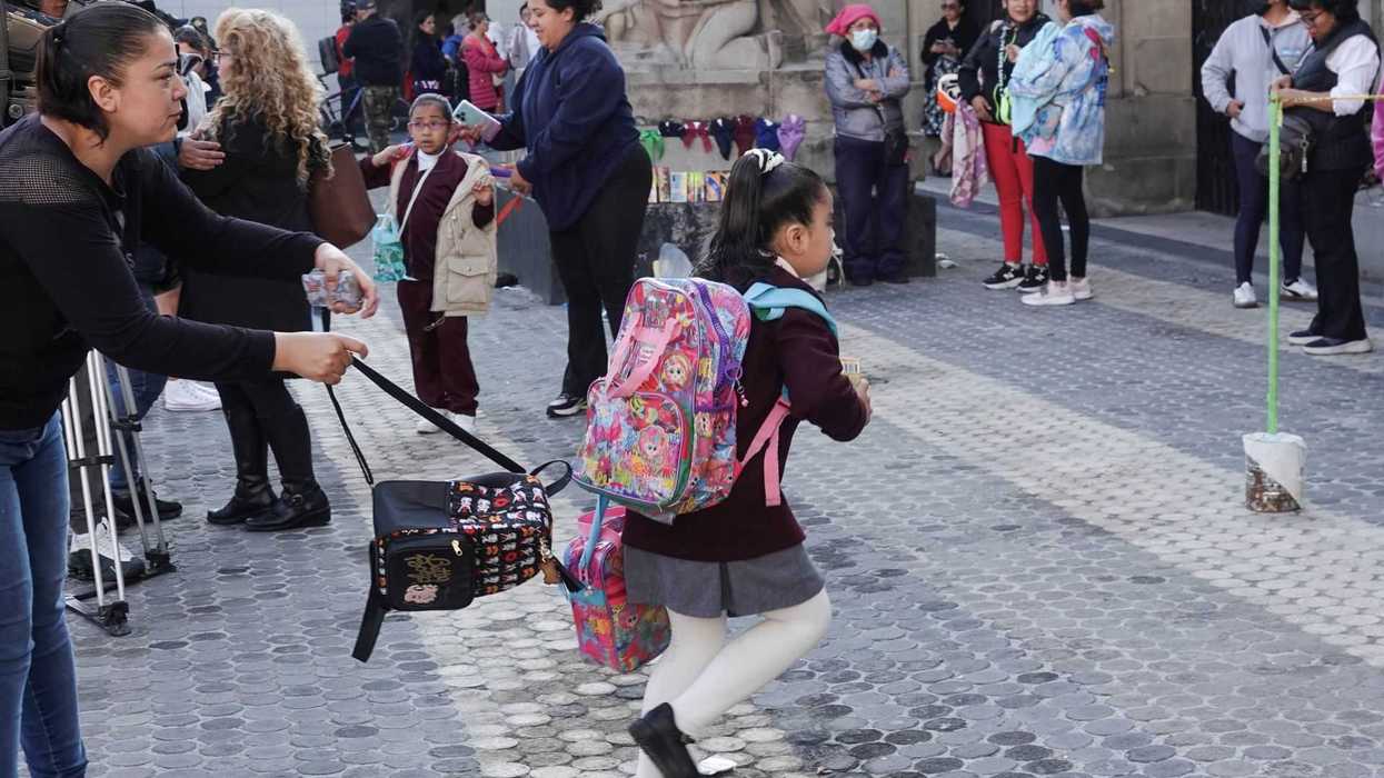 A woman helps a schoolgirl with backpacks in a busy outdoor setting.