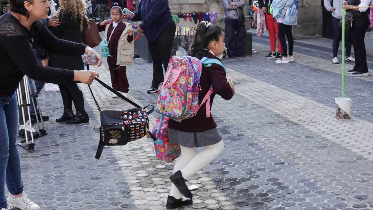 A woman helps a schoolgirl with backpacks in a busy outdoor setting.
