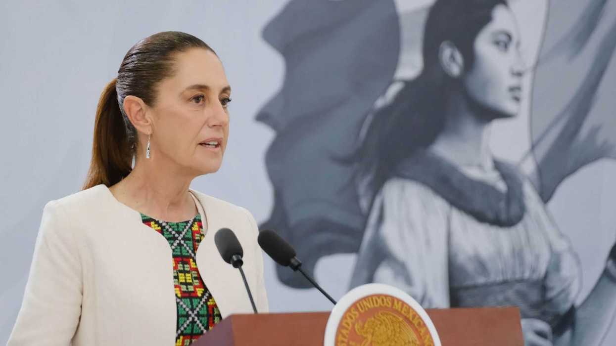 A woman speaks at a podium; a mural of a historical figure is in the background.