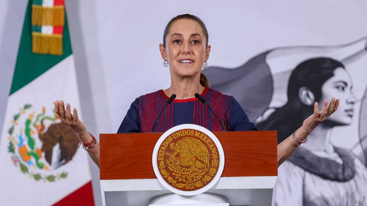 A woman speaks at a podium with a Mexican flag and mural in the background.
