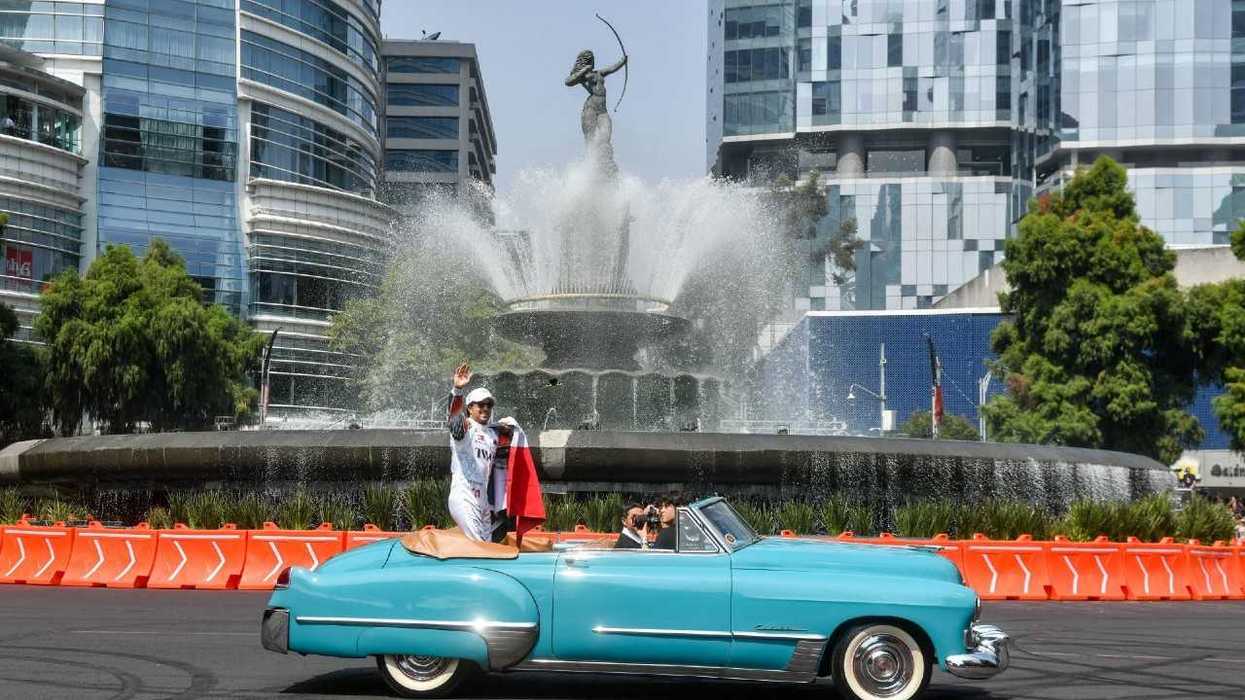 A woman waves from a turquoise vintage car passing a large fountain in a modern city plaza.