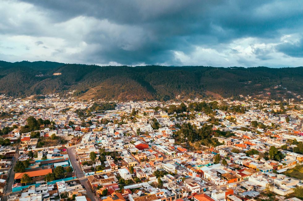 Aerial view of a town with colorful rooftops, surrounded by forested hills under a cloudy sky.