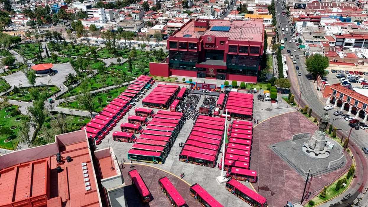 Aerial view of red buses parked in front of a large building, with a park nearby.
