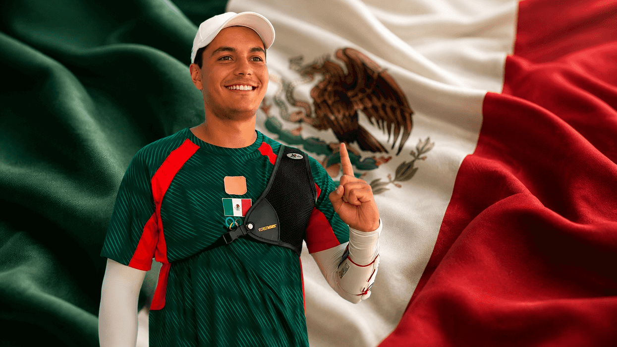Athlete in green jersey smiling, stands before the Mexican flag, raising his index finger.