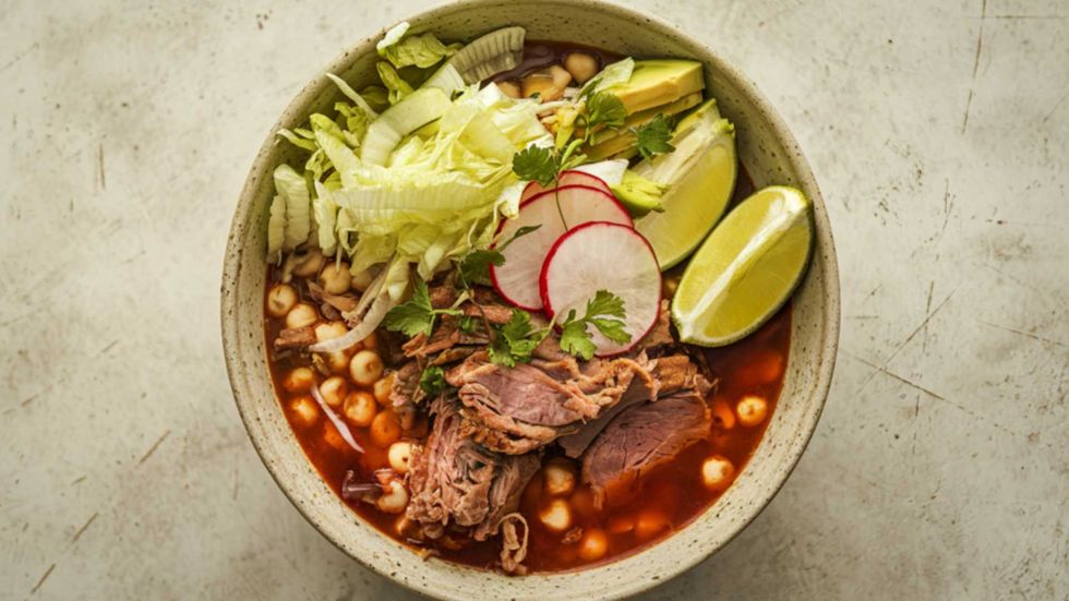 Bowl of pozole with pork, lettuce, radish, lime, avocado, and hominy.