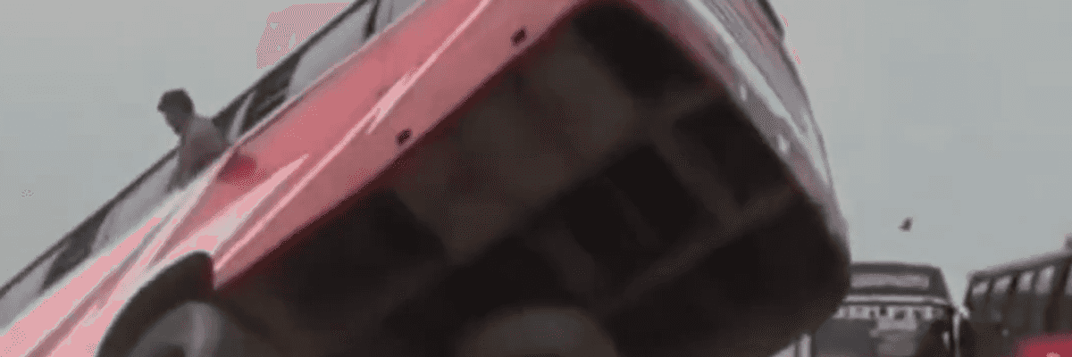 Bus tipping dangerously on a tilted platform, man standing beside.