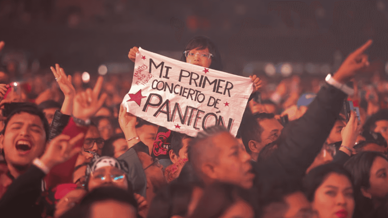 Child at concert holds a sign: "Mi primer concierto de Panteón", among a cheering crowd.