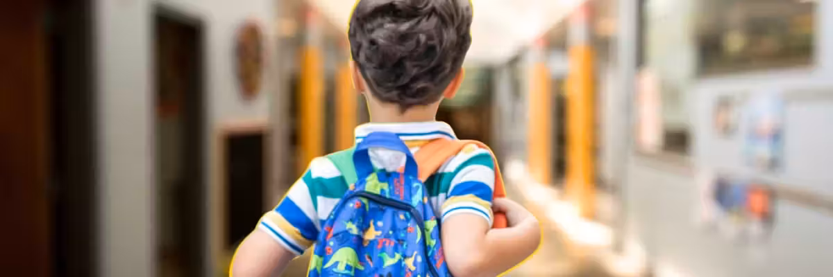 Child with dinosaur backpack in a bright hallway.