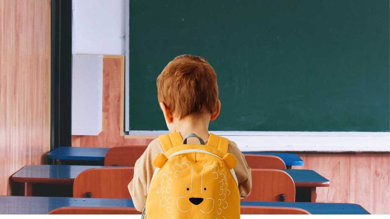 Child with lion backpack sits facing a chalkboard in a classroom.