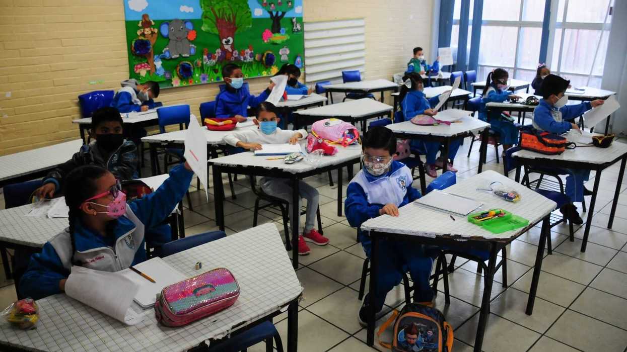 Children in a classroom wearing masks, seated at separate desks, holding papers.