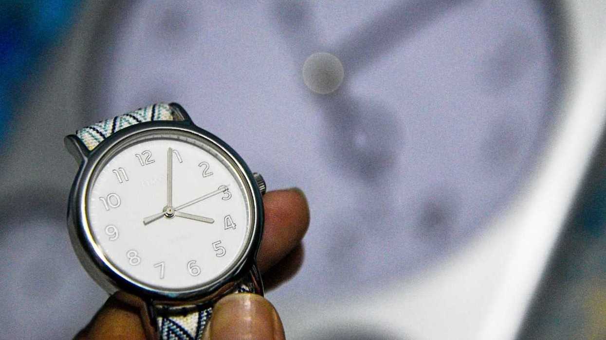 Close-up of a hand holding a wristwatch, with a blurred clock face in the background.