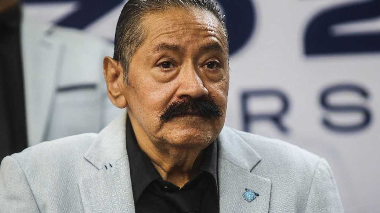 Close-up of a mustached man in a light gray blazer and black shirt at a press event.