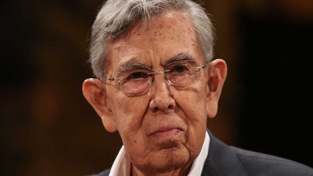 Close-up portrait of an older man with gray hair and glasses wearing a dark suit.