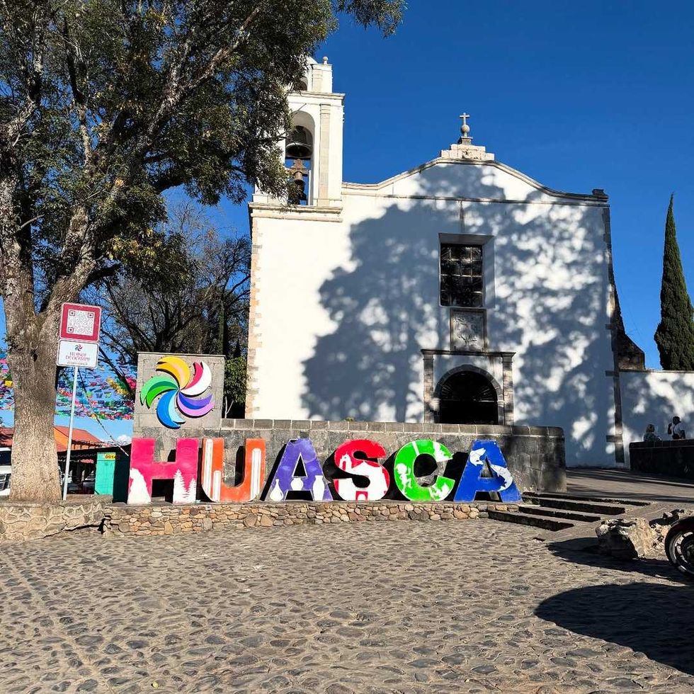 Colorful "HUASCA" letters in front of a white church under a clear blue sky.