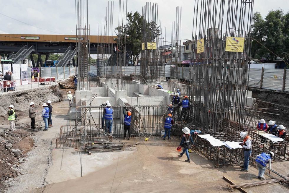 Construction site with workers, scaffolding, and rebar structures near an elevated walkway.