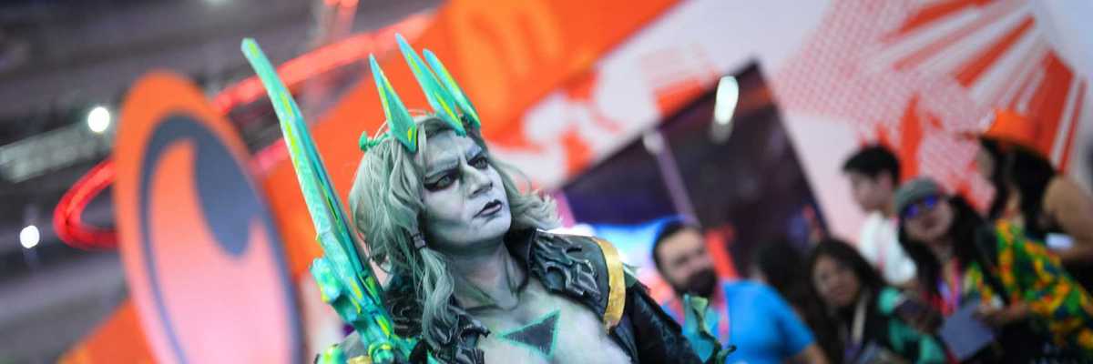 Cosplayer in elaborate armor with teal accents at a convention, orange backdrop.