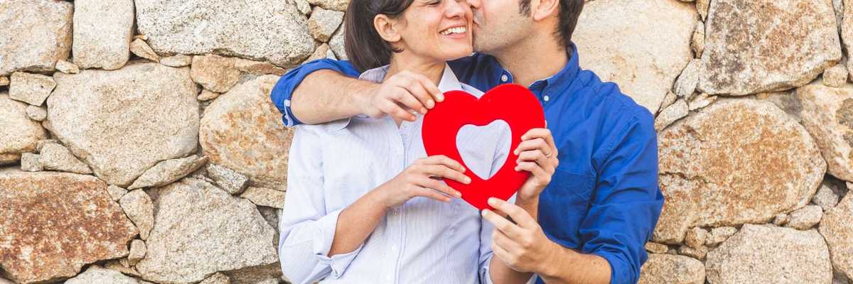 Couple holding a red heart decoration while standing against a stone wall.