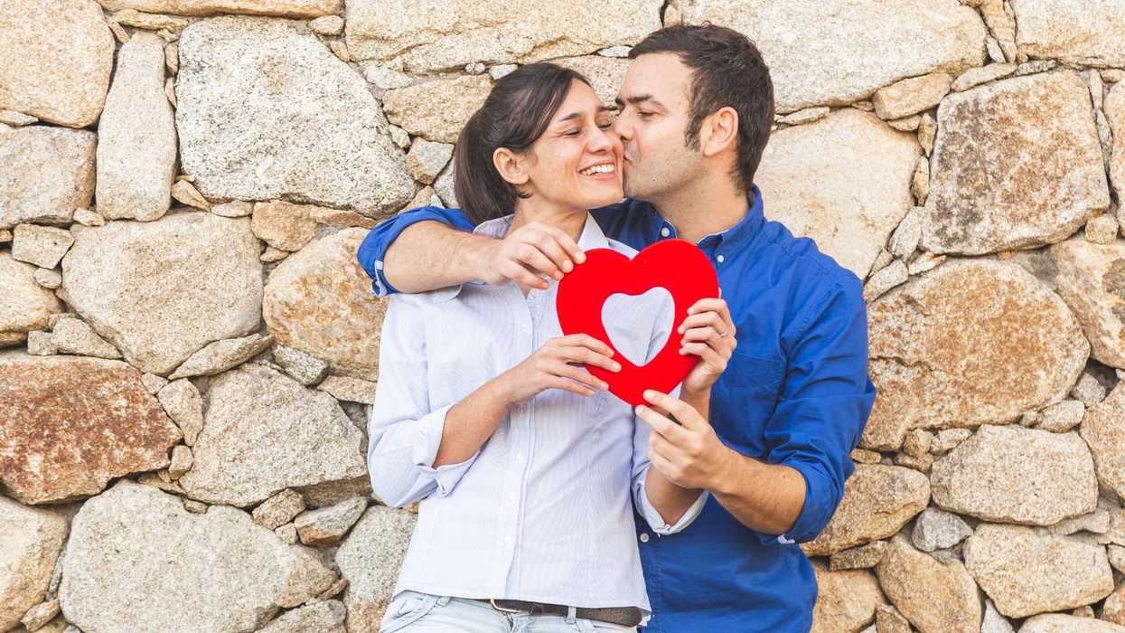 Couple holding a red heart decoration while standing against a stone wall.