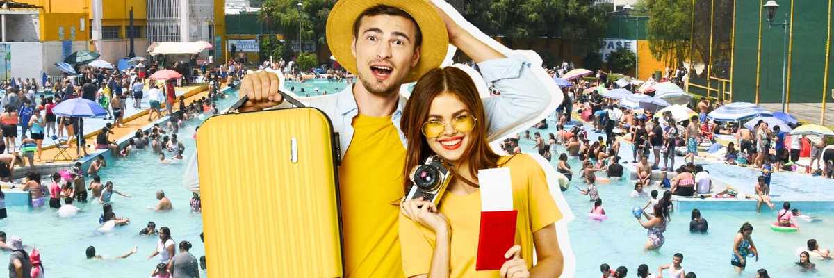Couple holding suitcase and camera at a crowded outdoor swimming pool.