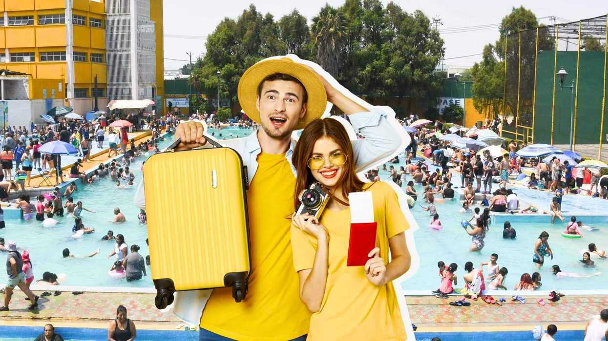Couple holding suitcase and camera at a crowded outdoor swimming pool.