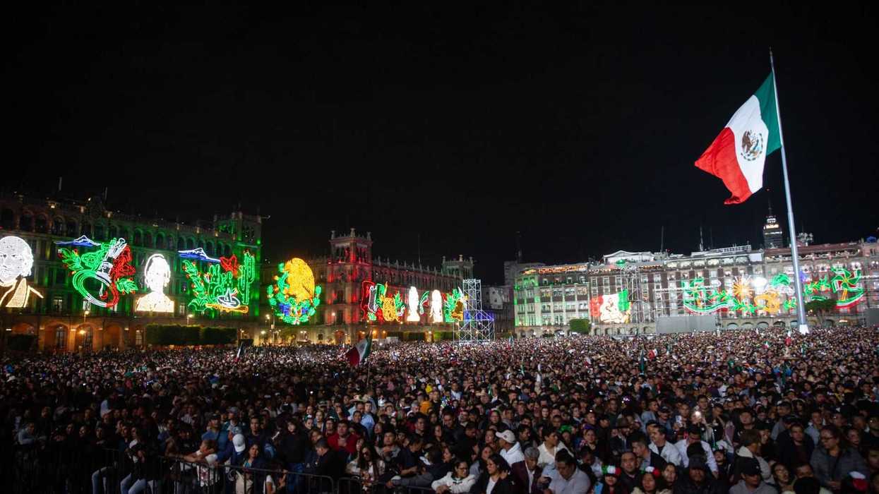 Crowd at night with colorful building lights and a large Mexican flag in the background.