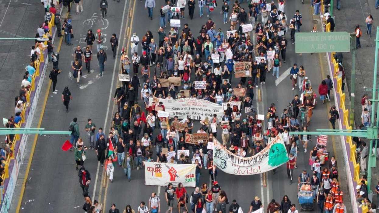 Crowd marching on a street, holding banners and signs, surrounded by onlookers.