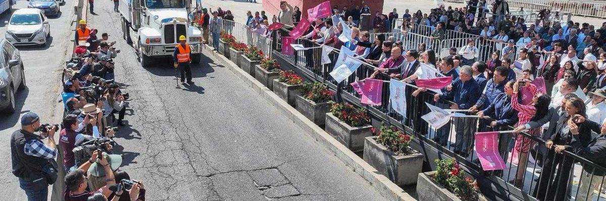 Crowd waves flags at truck on highway, photographers capture the scene in a busy urban area.