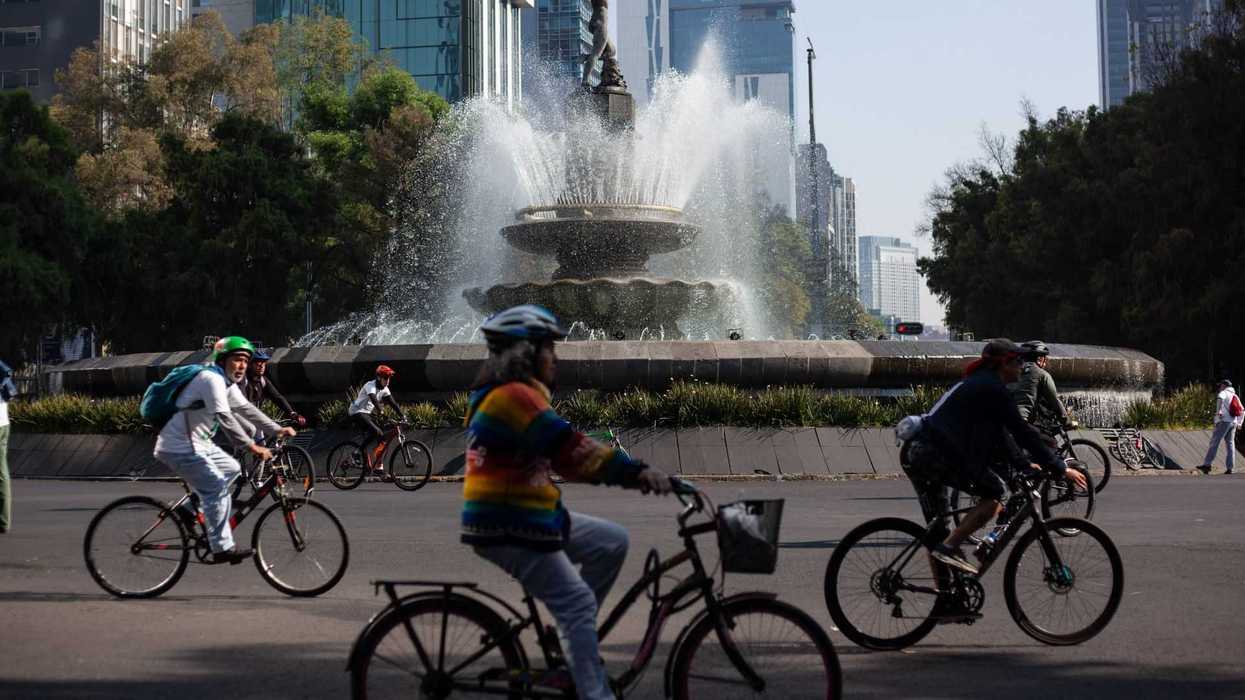 Cyclists ride past a large fountain in a bustling urban area with tall buildings.