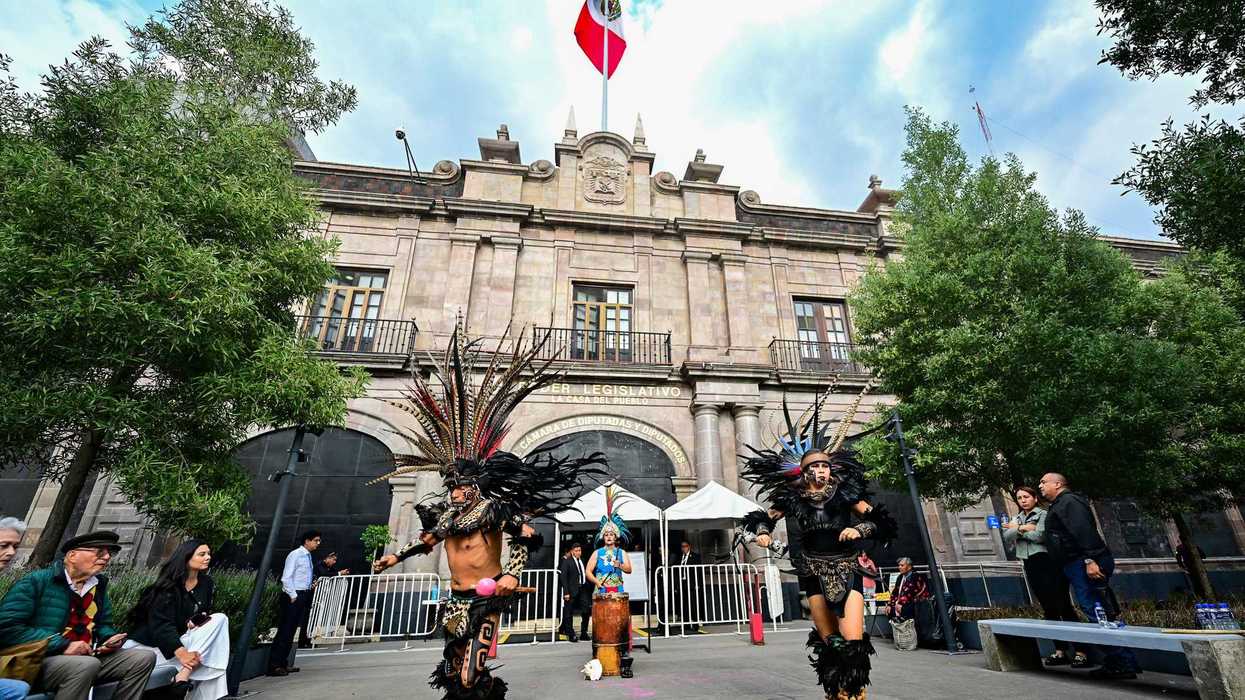 Dancers in traditional attire perform outside a historic building with a Mexican flag above.