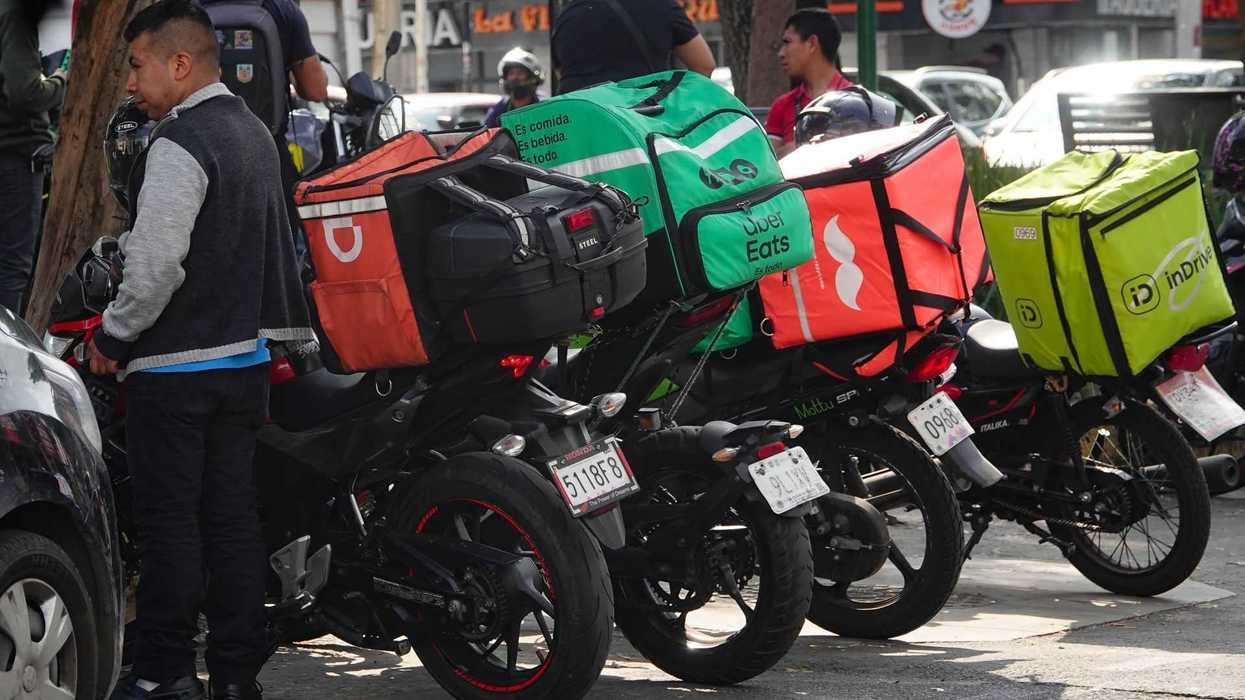 Delivery riders with branded backpacks gather by parked motorcycles.
