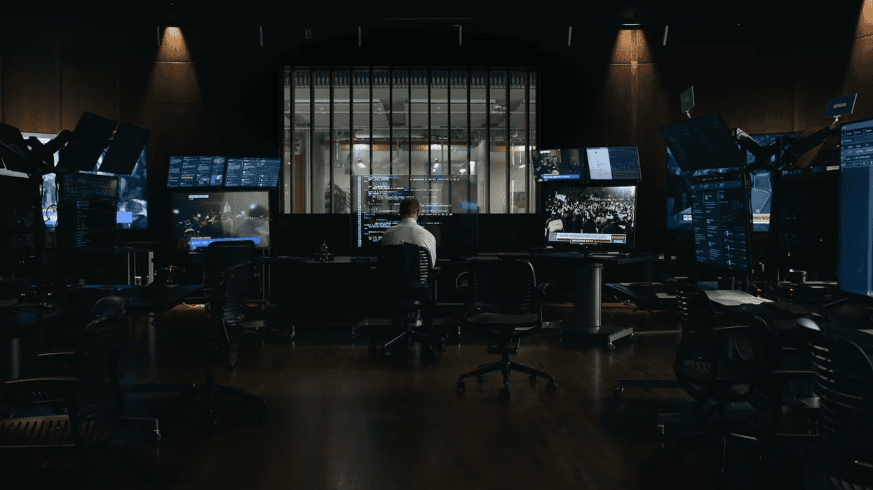 Dimly lit control room with multiple monitors and a person seated at the center.