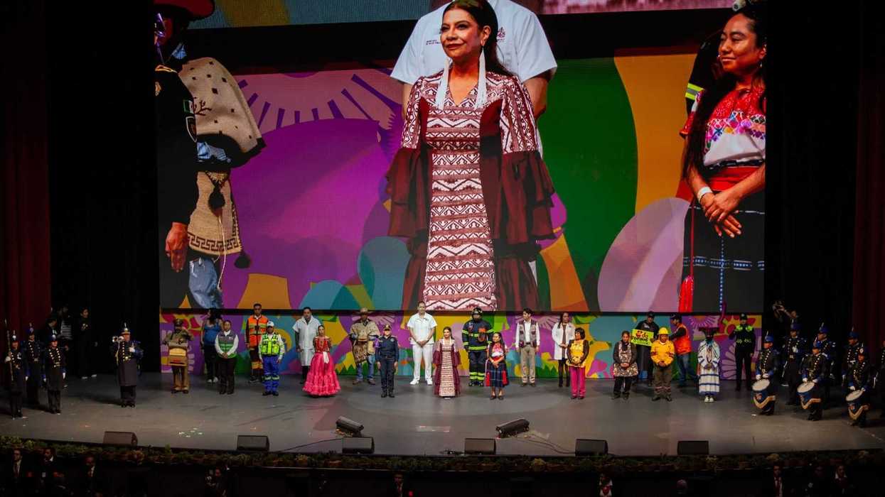 Diverse group in traditional attire stands on stage with large colorful backdrop display.