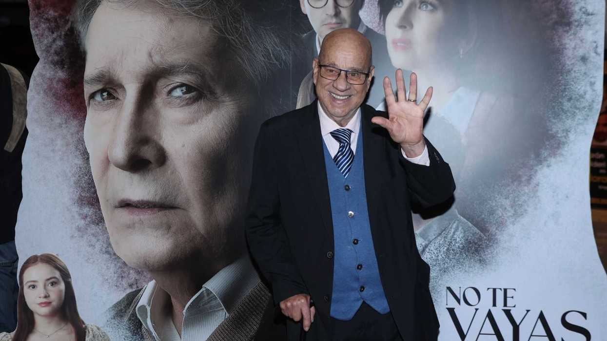 Elderly man waving, standing by a movie poster backdrop.
