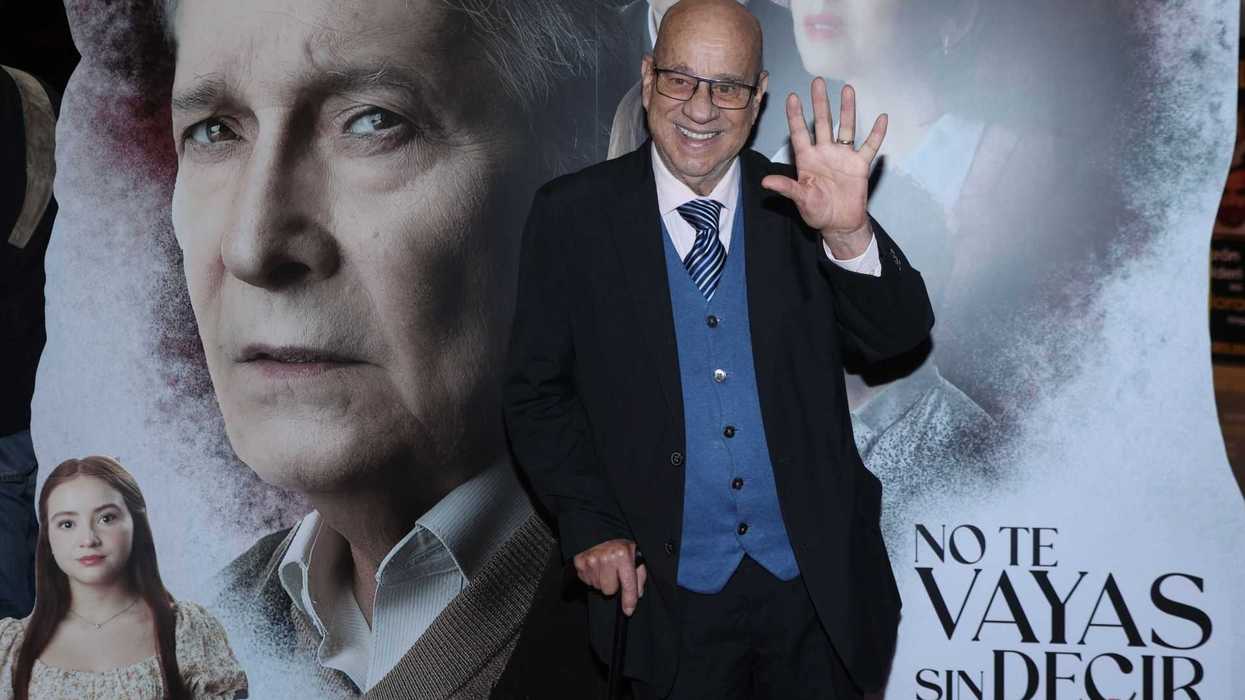 Elderly man waving, standing by a movie poster backdrop.