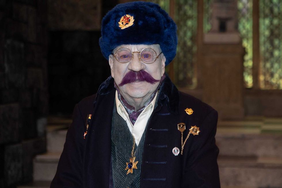 Elderly man with purple mustache wearing blue hat and decorated jacket indoors.