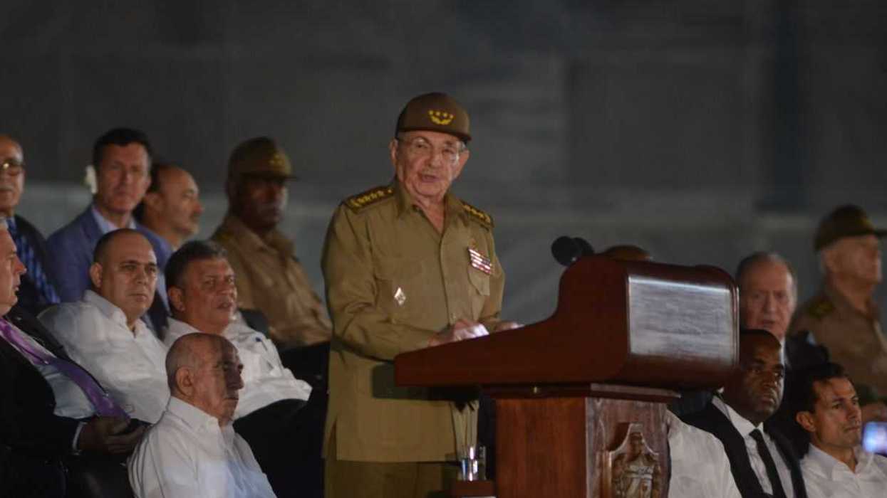 Elderly military officer speaking at podium, surrounded by seated officials.