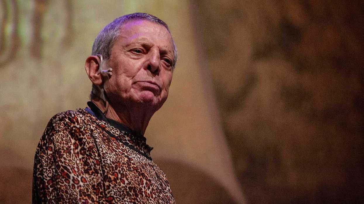 Elderly person on stage, wearing a leopard print shirt, with a microphone headset.