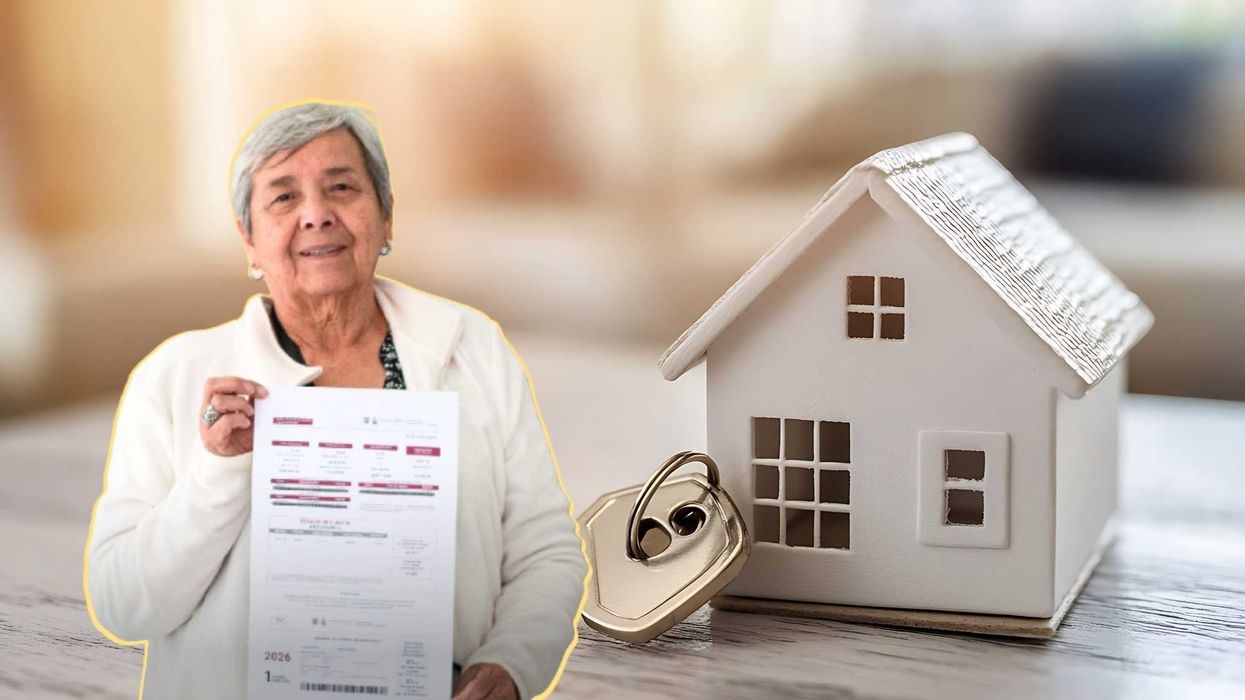 Elderly woman holding a document next to a miniature house and key on a table.