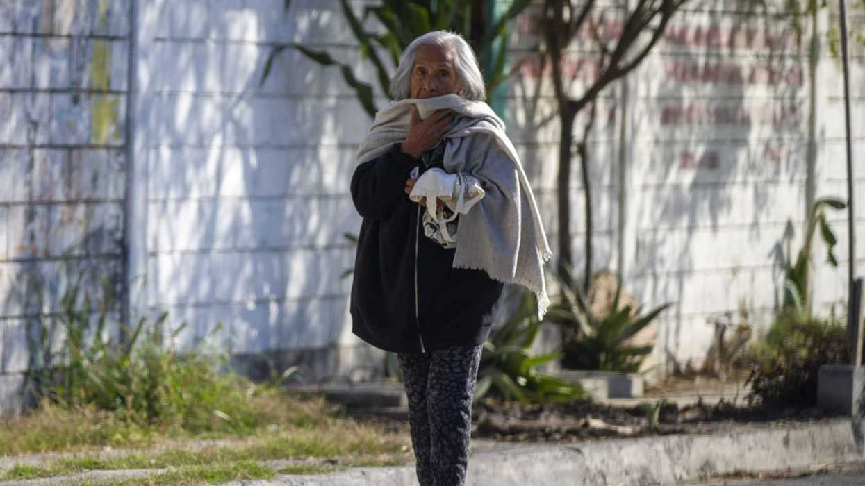 Elderly woman walking outdoors with a scarf, plants and a wall in the background.