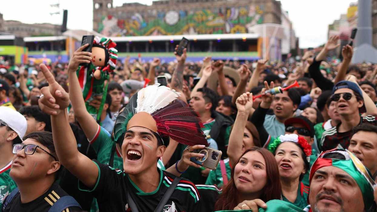 Excited crowd in festive attire cheering at outdoor event, holding flags and wearing team jerseys.