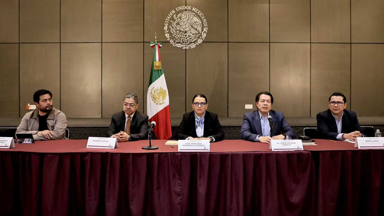 Five people at a conference table with a Mexican flag and emblem backdrop.