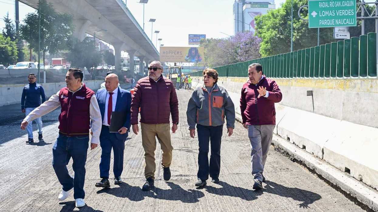 Five people walk on a construction site under a highway overpass.