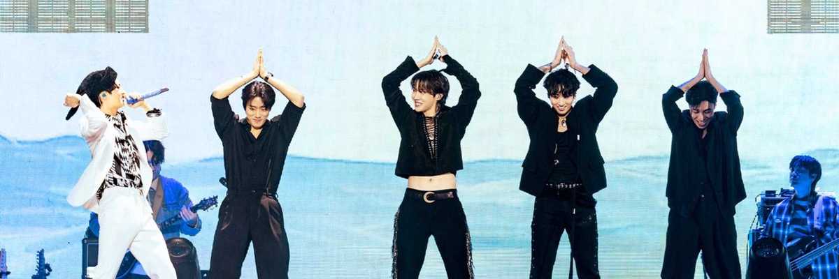 Five performers dance onstage, one singing with a microphone, colorful backdrop.